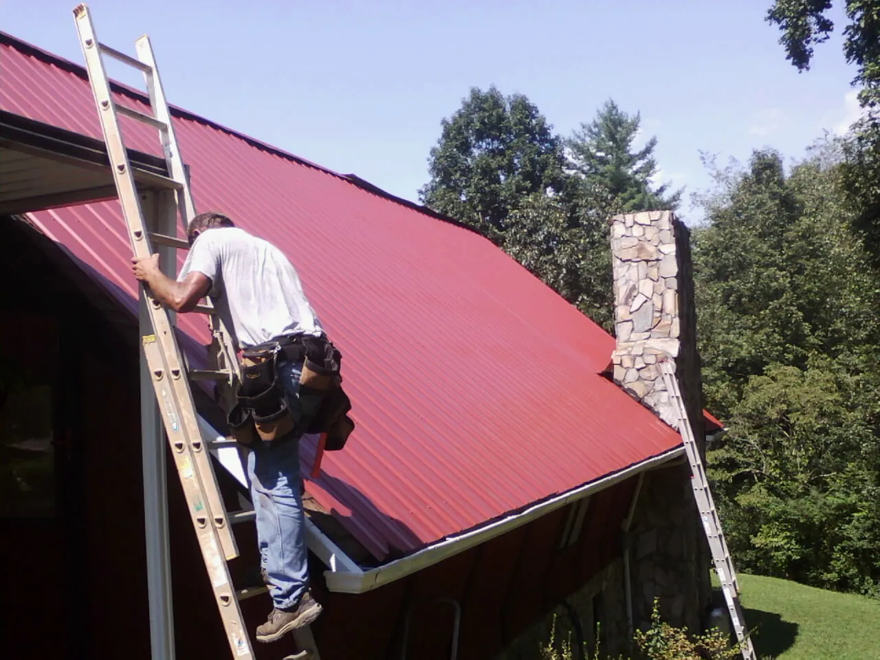 Roofer on a ladder beside a red metal roof and stone chimney — Soffit & Fascia Repair by Roofing Brothers in Saco